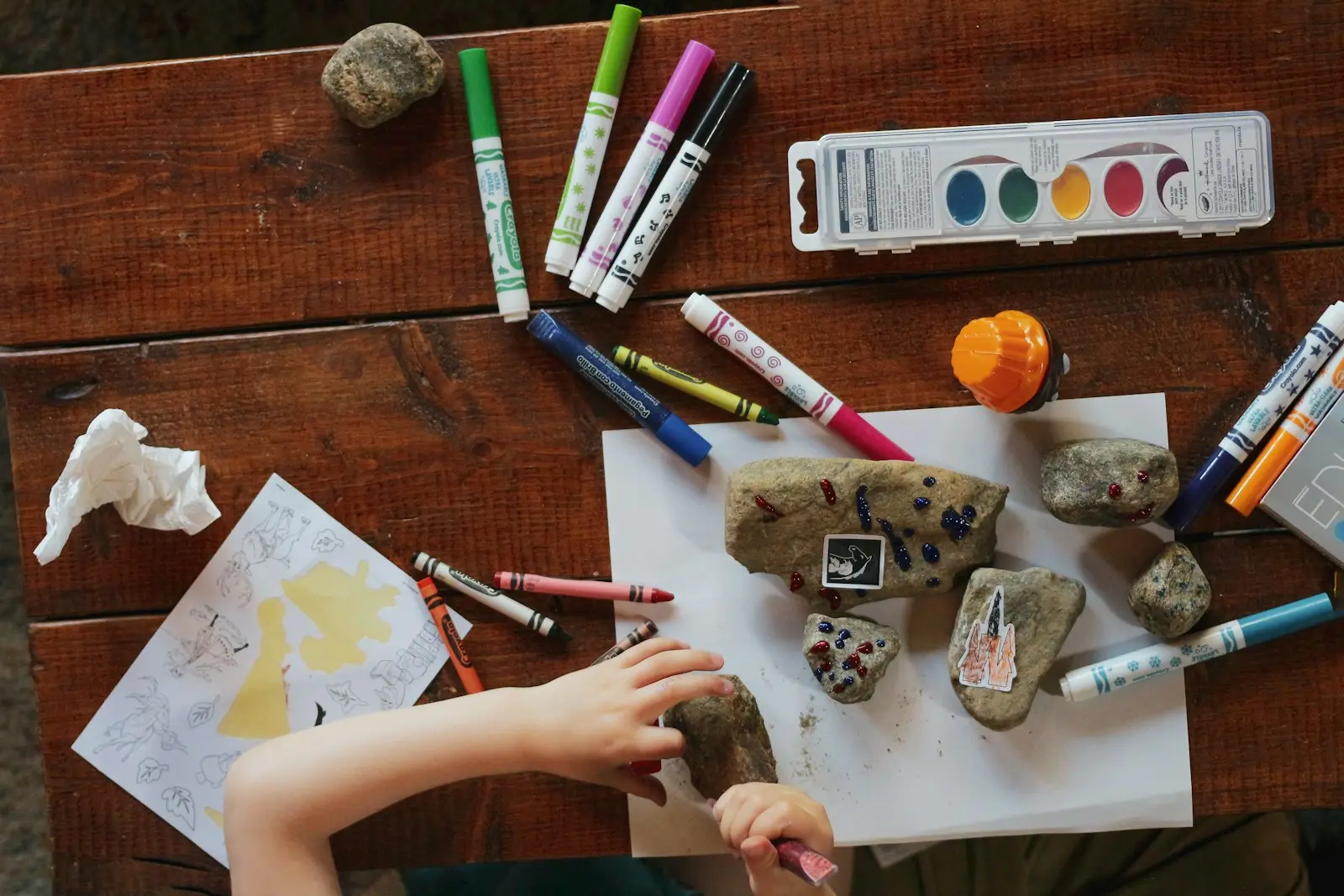A child drawing with markers beside painted rocks on a table